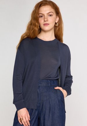 Cardigan blu navy con apertura frontale, polsini a coste e texture morbida, indossato sopra un top coordinato e abbinato a pantaloni in denim a vita alta.