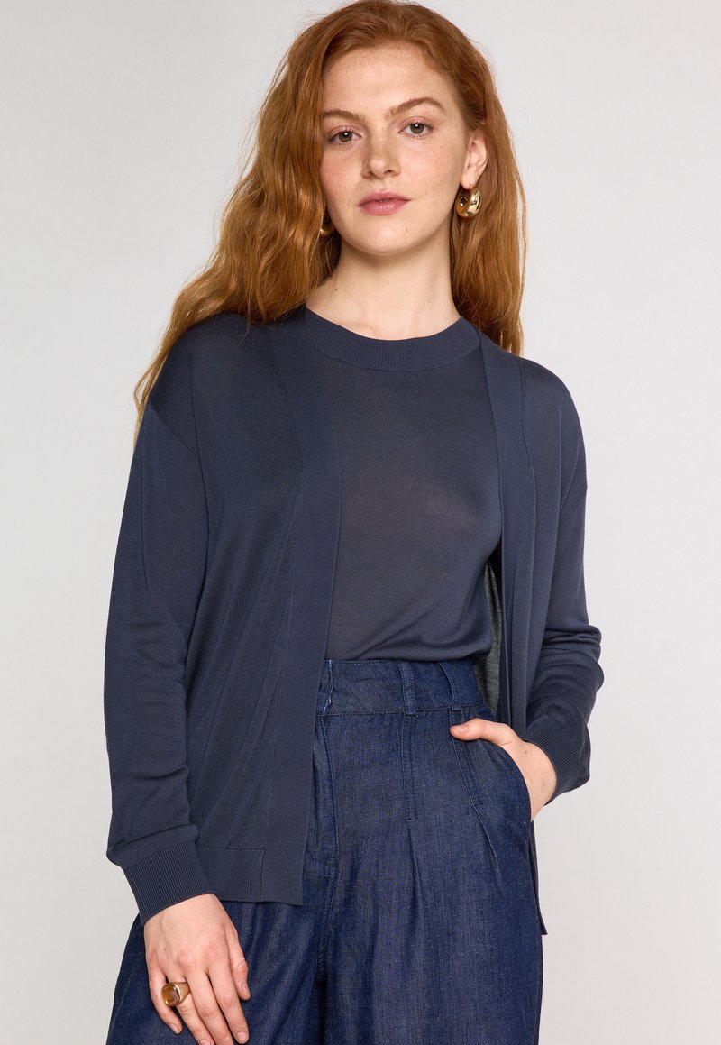 Cardigan marine avec devant ouvert, poignets côtelés et texture douce, porté par-dessus un top assorti et associé à un pantalon en denim taille haute.
