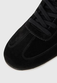 Punta di scarpa sneaker in suede nero con cuciture dettagliate e lacci neri, su sfondo grigio chiaro.