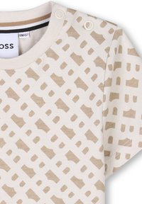 Chemise à manches courtes en coton léger, présentant un motif géométrique beige, encolure ronde avec deux boutons-pression sur une épaule.