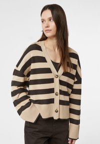 Beżowy kardigan z ciemnobrązowymi paskami, z dekoltem w serek, o oversize'owym kroju i trzema dużymi guzikami z przodu. Miękka dzianinowa faktura.