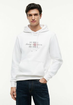 Mann trägt weißen Tommy Hilfiger Hoodie mit Kängurutasche und dunkelblauen Jeans, steht vor einfarbig weißem Hintergrund.
