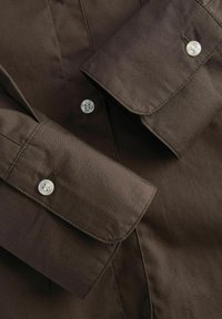 Camicetta marrone a maniche lunghe con una texture liscia, caratterizzata da polsini con bottoni e bottoni chiari, che enfatizzano un design classico.