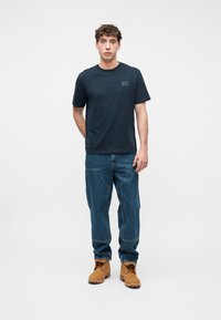 Νεαρός άνδρας που στέκεται με το πρόσωπο προς τα εμπρός, φορώντας μπλε ναυτικό μπλουζάκι, μπλε τζιν με μεγάλες τσέπες και καφέ μπότες με κορδόνια.