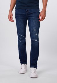 Jeans de mezclilla azul oscuro con desgastes y roturas visibles, diseño de pierna recta, combinados con zapatillas blancas y una camiseta navy.
