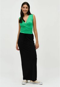 Groene wikkeltop met een diepe V-hals, gecombineerd met een lange zwarte rok met een elastische tailleband, gedragen met beige sneakers.
