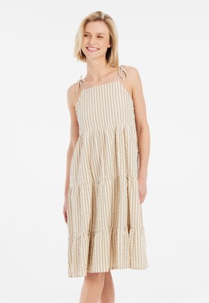 Vrouw lacht terwijl ze een beige-witte, verticale gestreepte mouwloze midijurk draagt met gebonden schouderbandjes tegen een effen achtergrond.