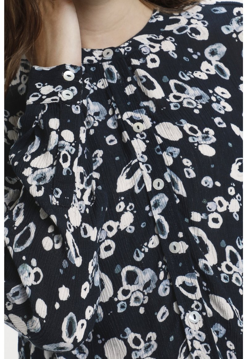 Svart- och vitt mönstrad skjorta med en texturerad tygkvalitet, med knappr detaljer vid ärmsluten och rund halsringning. Cirklar och detaljer i olika storlekar.