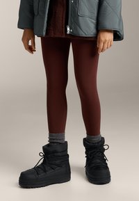 Czarne ocieplane buty na sznurowadła, bordowe legginsy i widoczny mankiet szarego swetra. Kurtka jest szara i pikowana.