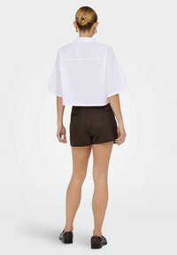 Kvinde med fletning i knolden iført en hvid cropped skjorte, mørkebrune shorts og mørke loafers, stående mod en ensfarvet lys baggrund.