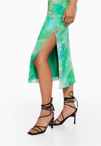 Robe tie-dye verte avec une fente sur le côté, fabriquée en tissu léger. Associée à des sandales noires à lanières avec un orteil carré et un talon.