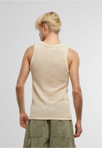 Beige, strikket tank topp med et mesh-mønster, som har brede armhull og rund halslinning. Brukt sammen med olivengrønne cargo-shorts.