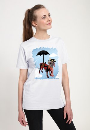 Kvinde iført hvid T-shirt med illustration af en hund i rødt og sort outfit, der holder en paraply, mod en blå himmel med skyer.
