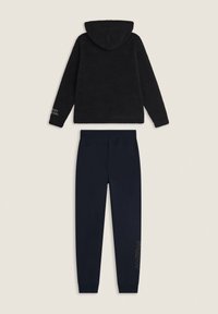 Giacca con cappuccio in pile nera realizzata in tessuto strutturato, abbinata a pantaloni morbidi blu navy con un dettaglio stampato sulla gamba sinistra.