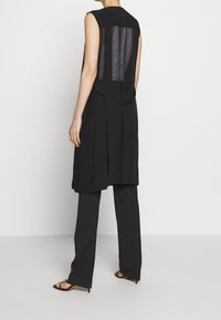 Svart ärmlös klänning med en genomskinlig rygg, kombinerad med lösa svarta byxor. Klänningen har en lagerdesign och mjuk textur.