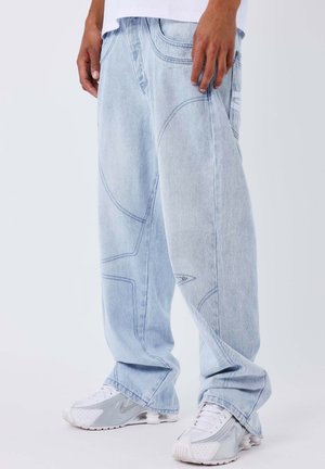 Lichtblauwe baggy jeans van denim, met contrasterende stikpatronen en een enkele zak met een reflecterende detail. Gecombineerd met witte sneakers.