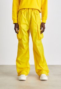 Pantaloni cargo giallo brillante con cintura elastica, molteplici tasche laterali e vestibilità rilassata, abbinati a sneakers bianche. Texture liscia.