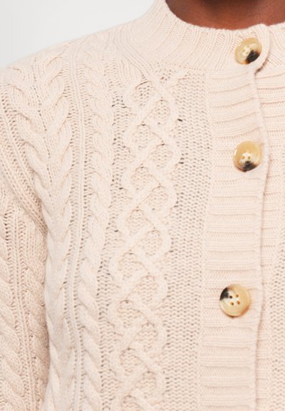 Knit cardigan in soft beige with textured cable patterns, featuring a ribbed neckline and three contrasting buttons on the front.
