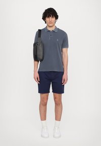 Polo grigio, pantaloni corti blu navy, sneakers bianche con suola spessa e una borsa a tracolla grigio scuro con zip e tracolla regolabile.