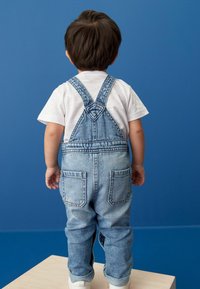 Lysblå denim overalls med justerbare stropper, to baglommer og opsmøgede ben, båret med en hvid t-shirt, foran en blå baggrund.