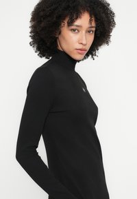 Črna rebrasta dolgotrajna majica z visokim ovratnikom, prilegajočo se silhueto in majhno podrobnostjo logotipa na prsih. Gladek tekstura, minimalističen dizajn.
