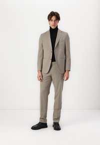 Béžový vzorovaný oblek s prispôsobeným sakom a nohavicami; skombinovaný s čiernym rolákom. Čierne topánky dopĺňajú outfit.