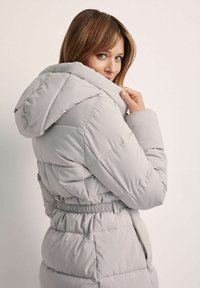 Jasnoszara pikowana kurtka z kapturem, elastycznym paskiem w talii i pikowanymi detalami. Zawiera boczne kieszenie i elegancki design.