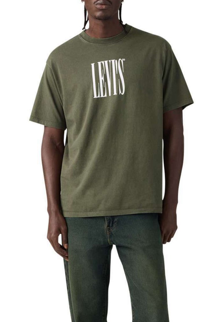 Hombre con camiseta Levi's color verde oliva y vaqueros verde oscuro, de pie contra un fondo blanco.
