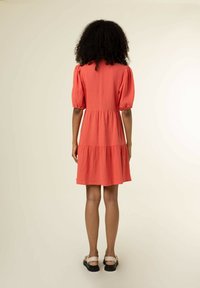 Korallenfarbenes, schichtetes Kleid aus leichtem Stoff, mit Ballonärmeln und strukturiertem Muster. Ergänzt durch flache Sandalen.