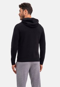 Schwarzer, gestrickter Hoodie mit lockerem Sitz, der eine weiche Textur und eine große, lockere Kapuze aufweist. Kombiniert mit grauen Jogginghosen.