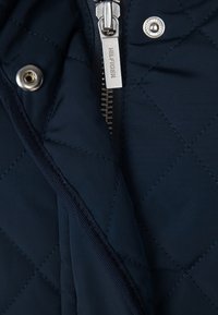 Granatowa pikowana kurtka, detal z srebrnym zamkiem błyskawicznym z logo, zatrzaskiem oraz kontrastowym przeszyciem wzdłuż krawędzi.