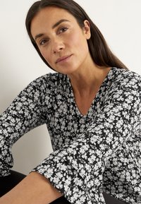 Kvinna med långt mörkt hår som bär en svart- och vitblommig långärmad topp, sitter och ser direkt in i kameran.