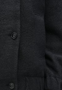Μαύρο παλτό από μαλλί με υφή, δύο μπροστινές τσέπες και κλείσιμο με κουμπί. Ο ιστός φαίνεται παχύς και ζεστός.