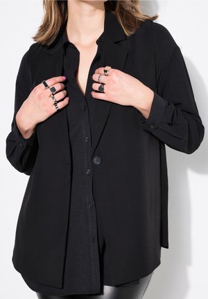 Femme portant une chemise noire boutonnée et un blazer, montrant plusieurs bagues sur les deux mains sur un fond blanc.