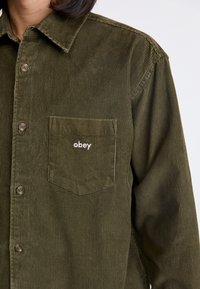 Olive zelena kordna srajca z gumbi na sprednji strani, žeponastavljen pokrovček in izvezenim logotipom "obey". Ima vertikalno rebrasto teksturo in nazaj usmerjen ovratnik.