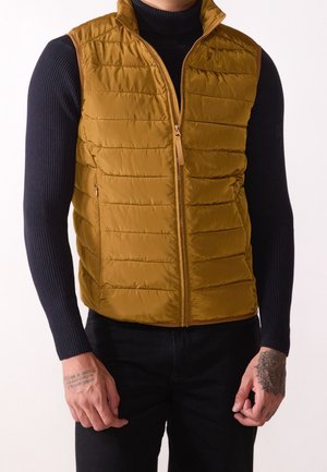 Homme portant une veste matelassée jaune moutarde par-dessus une chemise noire à manches longues côtelée et un pantalon noir, montrant des mains tatouées.