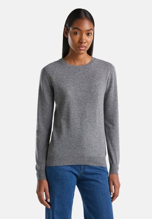 Femme aux longs cheveux raides portant un pull gris uni à manches longues et un jean bleu, sur un fond blanc.