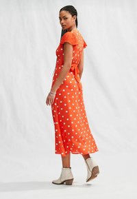 Abito lungo arancione con pois bianchi, maniche a sbuffo corte, dettaglio con lacci in vita, indossato con stivaletti color crema chiaro.
