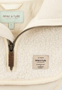 Cremefarbener Fleece-Pullover mit Reißverschlusskragen, der eine strukturierte Oberfläche, einen Leder-Reißverschlussanhänger und ein Etikett mit der Marke und dem Herstellungsort aufweist.