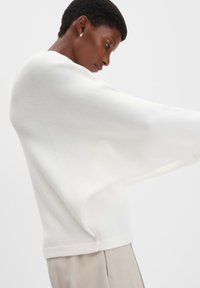Maglione bianco a maglia con maniche ampie e morbide e una texture liscia, indossato sopra pantaloni beige su misura. Design minimale senza motivi visibili.
