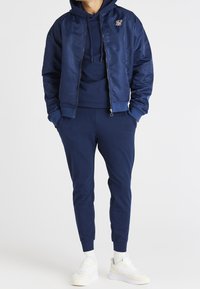 Marinblå bomberjacka med glansig finish, kombinerad med matchande hoodie och joggers. Har sidofickor och logodetaljer. Vita sneakers.