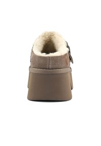 Bruine suède instapschoen met een fluffy witte voering, vlakke rubbere zool en een ronde achterkant. Inclusief een decoratieve band aan de zijkant.