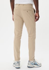 Beige trousers with a straight cut, featuring back pockets with button closures. Worn with gray and blue athletic shoes.