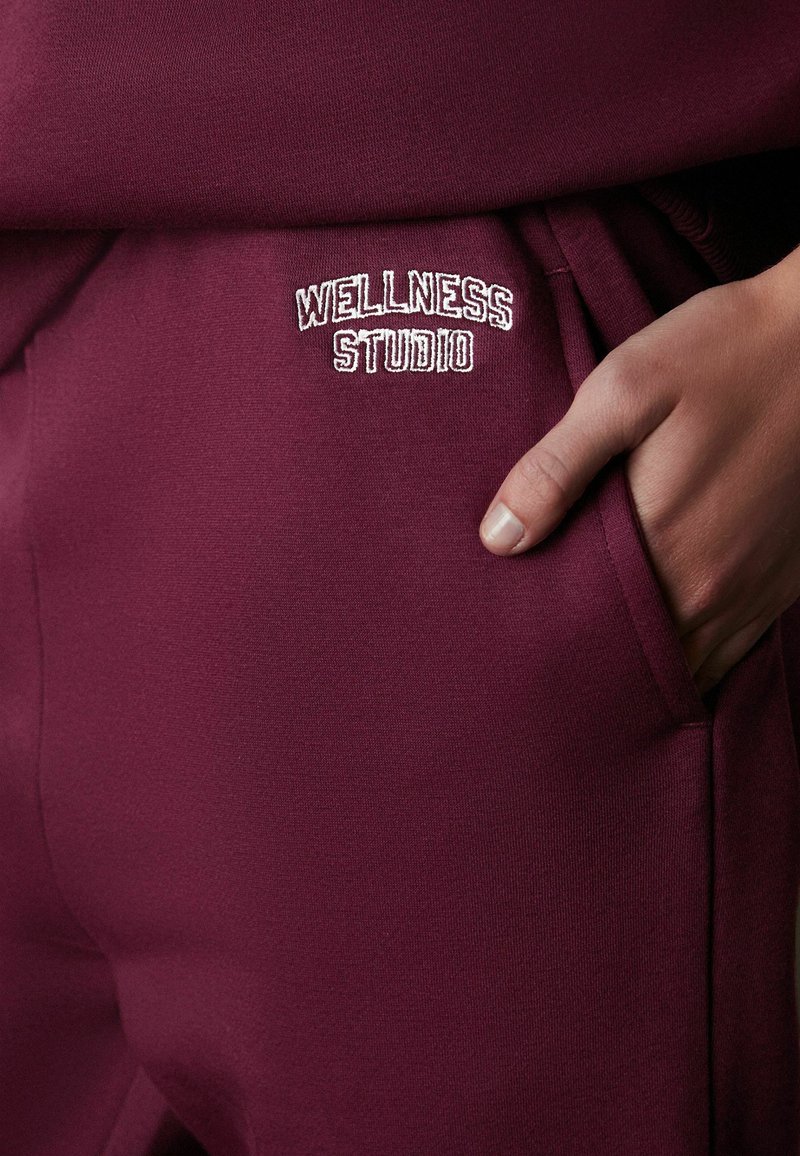 Burgundski džemperi s glatkom teksturom i izvezenim logom "WELLNESS STUDIO" u bijeloj boji na kuku, sa bočnim džepovima i sportskim dizajnom.