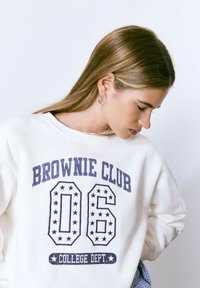 Sudadera blanca con la inscripción "BROWNIE CLUB 06" y "COLLEGE DEPT." en letras azul marino y patrones de estrellas. Material suave y corte relajado.