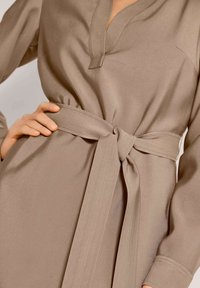 Beige langemouw jurk met een v-hals, strikceintuur op de taille en een soepele stof met een subtiele glans. Let op de gerimpelde mouwdetails.