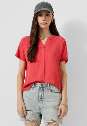 Vrouw draagt een rode blouse met korte mouwen, lichtgrijze gescheurde denimshorts, een grijze pet, gouden ronde oorbellen en een gouden armband, staand tegen een effen achtergrond.