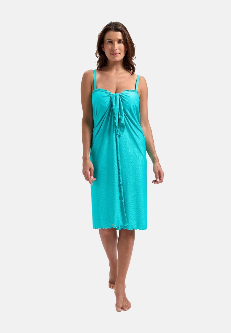 Vestido sin mangas turquesa con un lazo en la parte delantera, tejido texturizado y dobladillo con forma de concha. La modelo está descalza frente a un fondo sencillo.