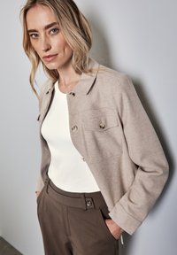 Beige uldblandingsjakke med sidelommer, sølvknapper og krave, parret med en hvid top og brune skræddersyede bukser. Glat tekstur.