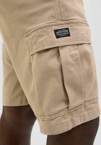 Beżowe bawełniane szorty cargo z kieszeniami bocznymi i przeszyciami. Metka na kieszeni informuje "Jack & Jones." Tekstura wydaje się gładka i lekka.